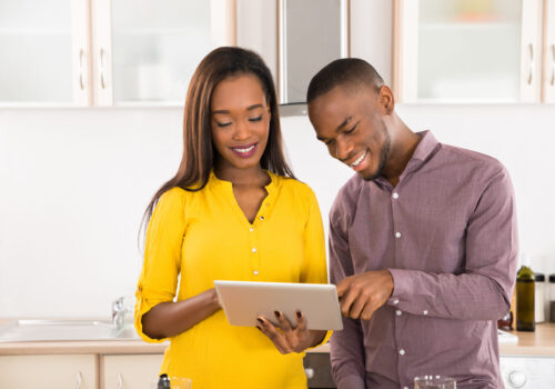 Happy,Young,Couple,Standing,In,Kitchen,Using,Digital,Tablet,With
